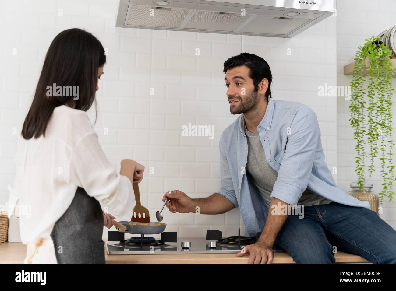 Ein lächelnder Mann sitzt auf der Küchentheke und spricht mit einer Frau, die auf dem Gasherd kocht. Ein Paar genießt zwanglose Momente in der Küche. Moderner Lebensstil, d Stockfoto