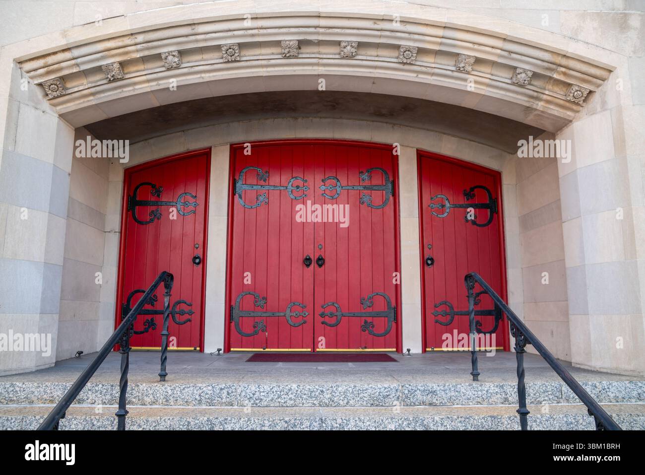 Ein markantes Paar roter Doppeltüren in einer strukturierten beigefarbenen Wand, die unter warmem Nachmittagslicht dramatische Schatten zaubern, Symmetrie und Vintage-Charme für den Bogen Stockfoto