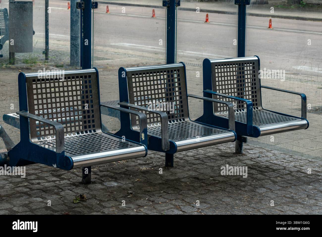 Drei moderne Metallbänke befinden sich an einer Bushaltestelle in einem Stadtgebiet. Die Sitzbänke haben ein Gitterdesign und sind auf einer gepflasterten Fläche aufgestellt, die für wartende Passagiere bereit ist. Stockfoto
