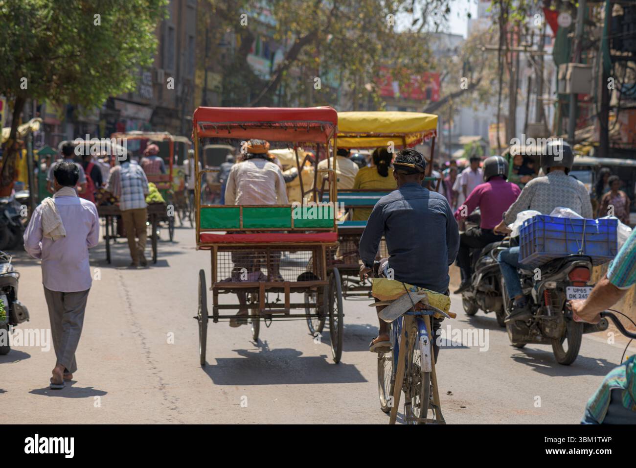 Indien, Varanasi, 16. März 2025. Zwei Männer schieben mitten in der Altstadt von Varanasi einen schwer beladenen Handwagen über eine geschäftige Straße, die von Geschäften und Verkehr gesäumt ist Stockfoto