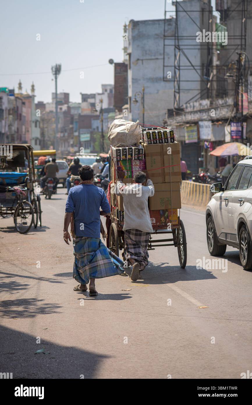 Indien, Varanasi, 16. März 2025. Zwei Männer schieben mitten in der Altstadt von Varanasi einen schwer beladenen Handwagen über eine geschäftige Straße, die von Geschäften und Verkehr gesäumt ist Stockfoto