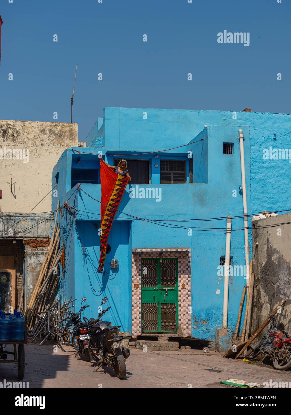 Indien, Varanasi, 16. März 2025. Frau hängt leuchtend rote Stoffe an einer hellblauen Hausfassade in einer engen Varanasi-Gasse unter einem klaren blauen Himmel. Stockfoto