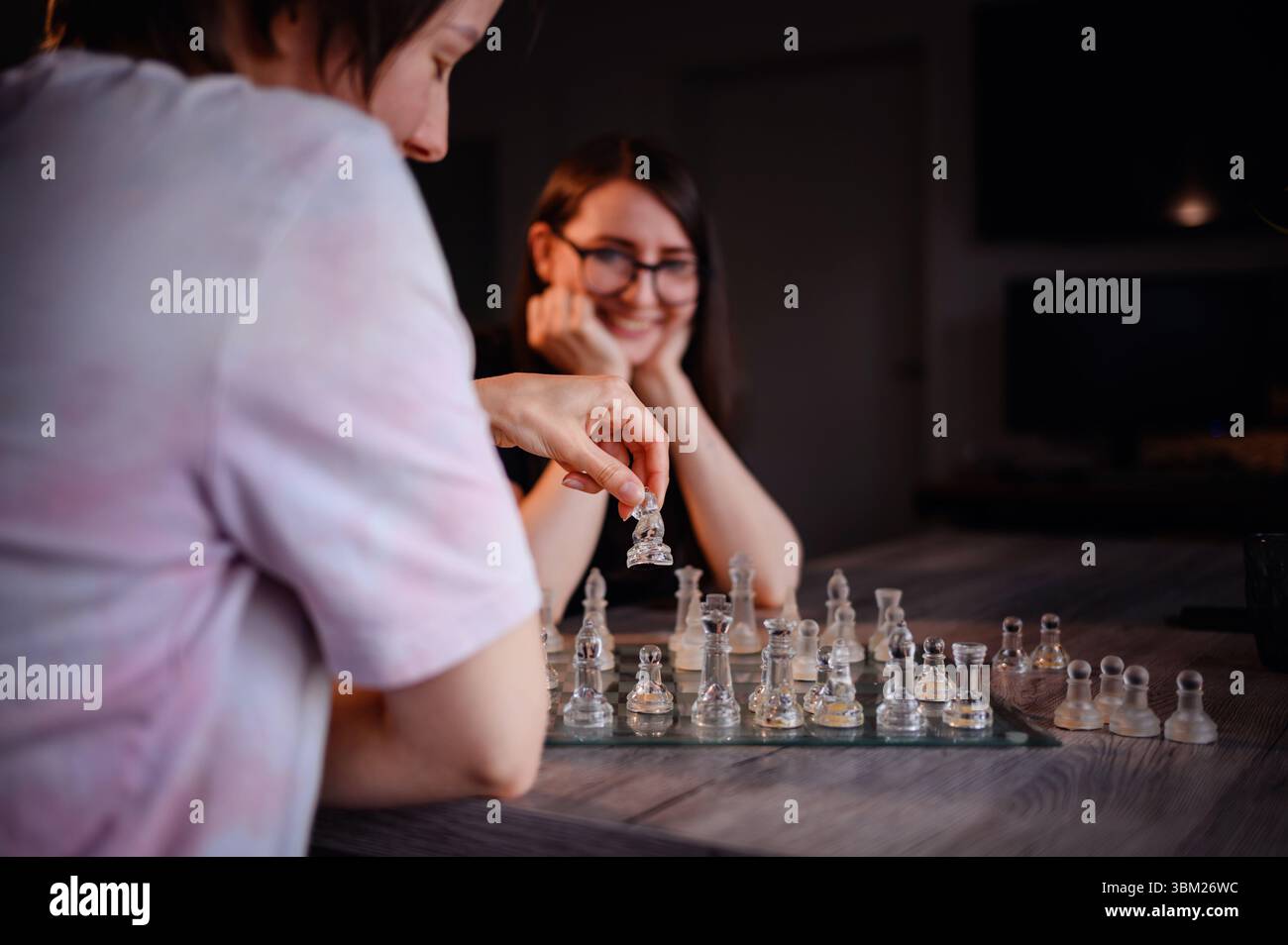 Zwei Freunde teilen sich während eines Schachspiels einen lustigen Moment, einer lächelt, der andere bewegt sich mit einem Glasstück an einem Holztisch. Stockfoto