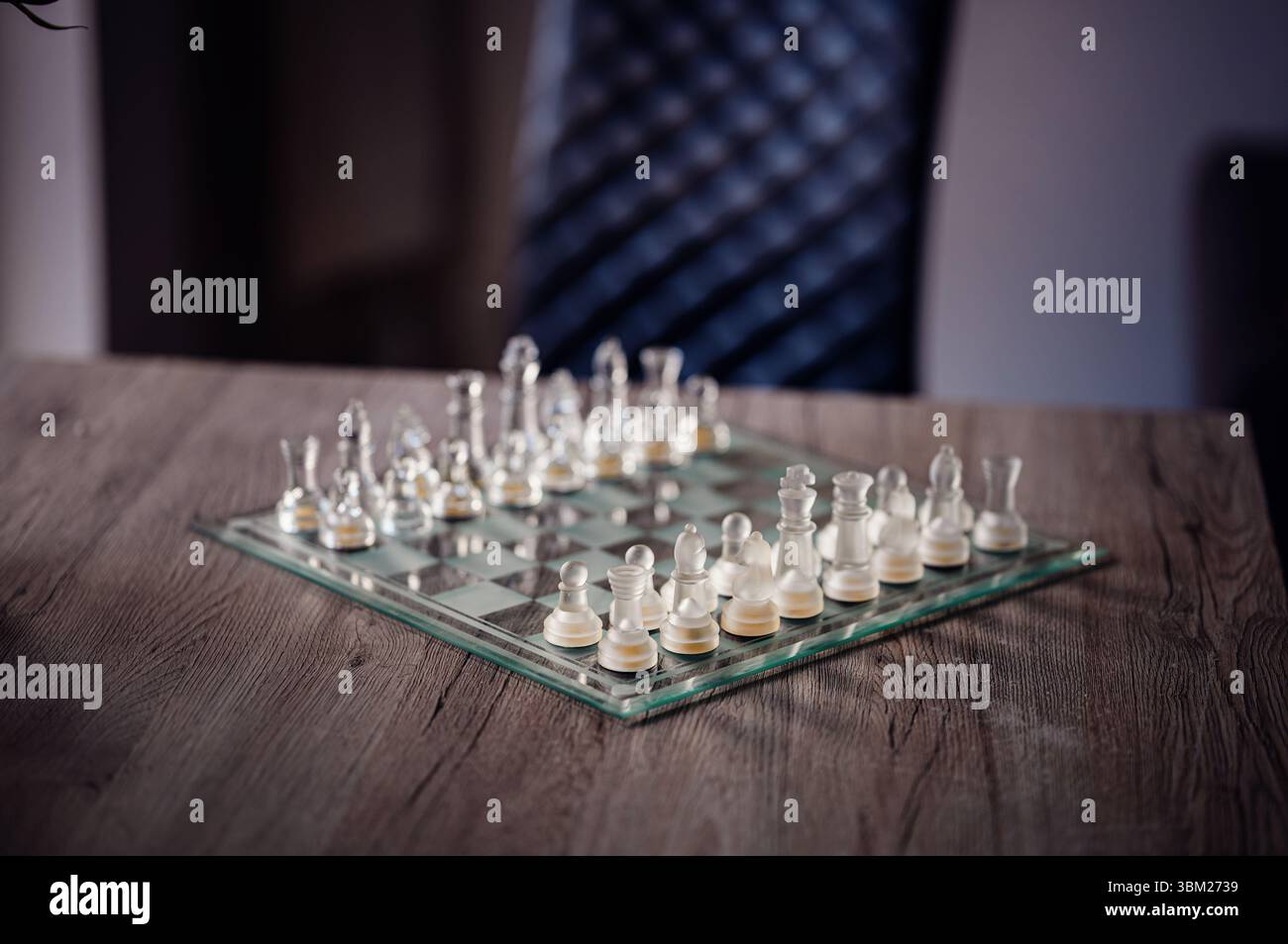 Elegante Schachstücke aus Glas, die auf einem transparenten Brett auf einem Holztisch angeordnet sind, mit einem verschwommenen Stuhl im Hintergrund. Stockfoto