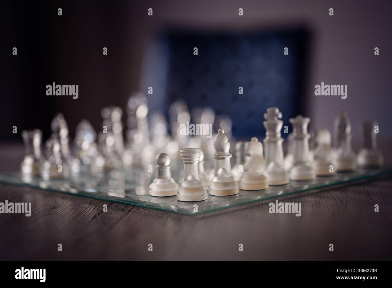 Elegante Schachstücke aus Glas, die auf einem transparenten Brett auf einem Holztisch angeordnet sind, mit einem verschwommenen Stuhl im Hintergrund. Stockfoto