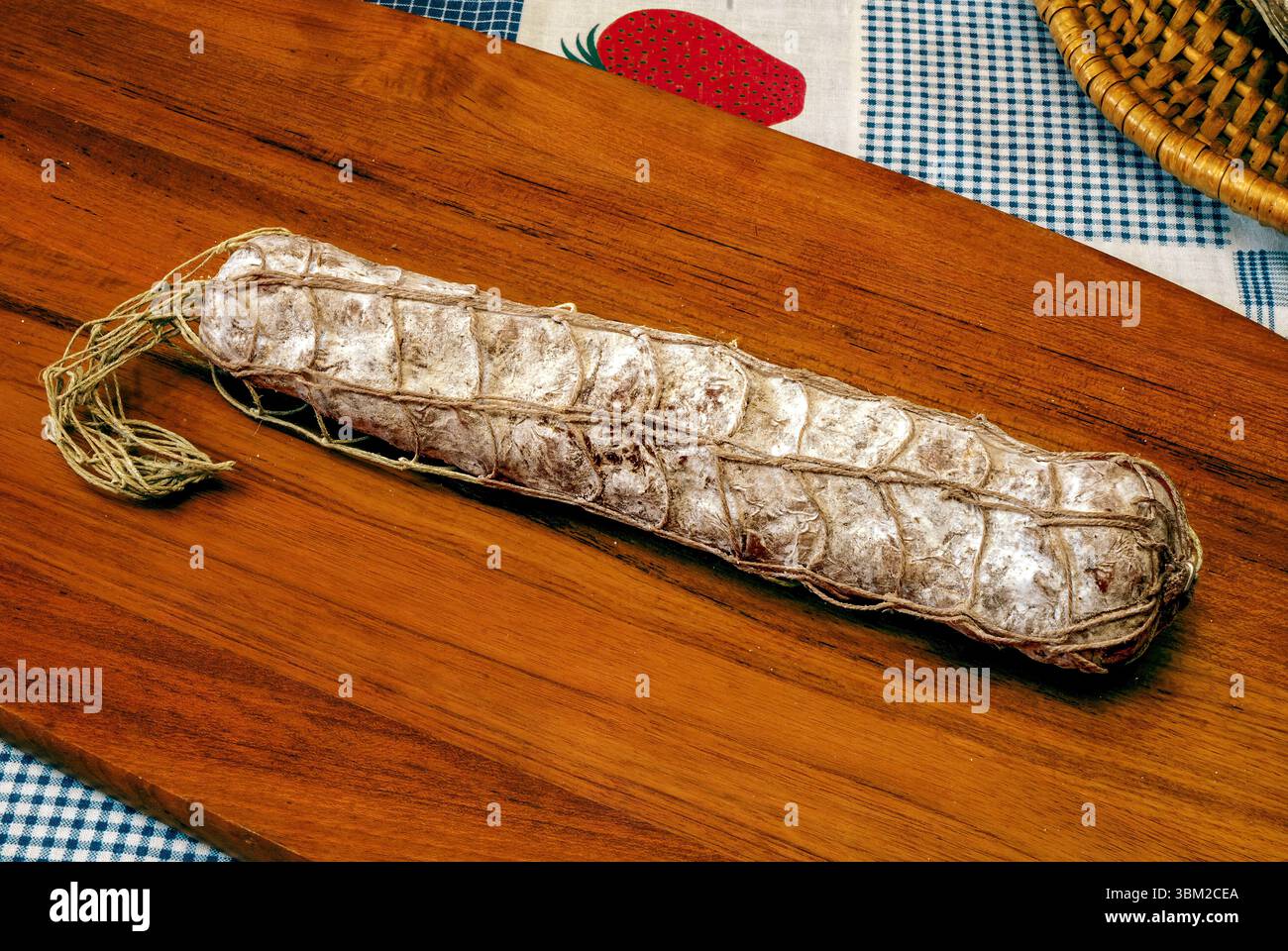 Die in ein Netz eingefasste Wurst liegt auf einem hölzernen Schneidebrett, auf einer karierten Tischdecke, mit einer Korbkante und einer stilisierten Erdbeere Stockfoto