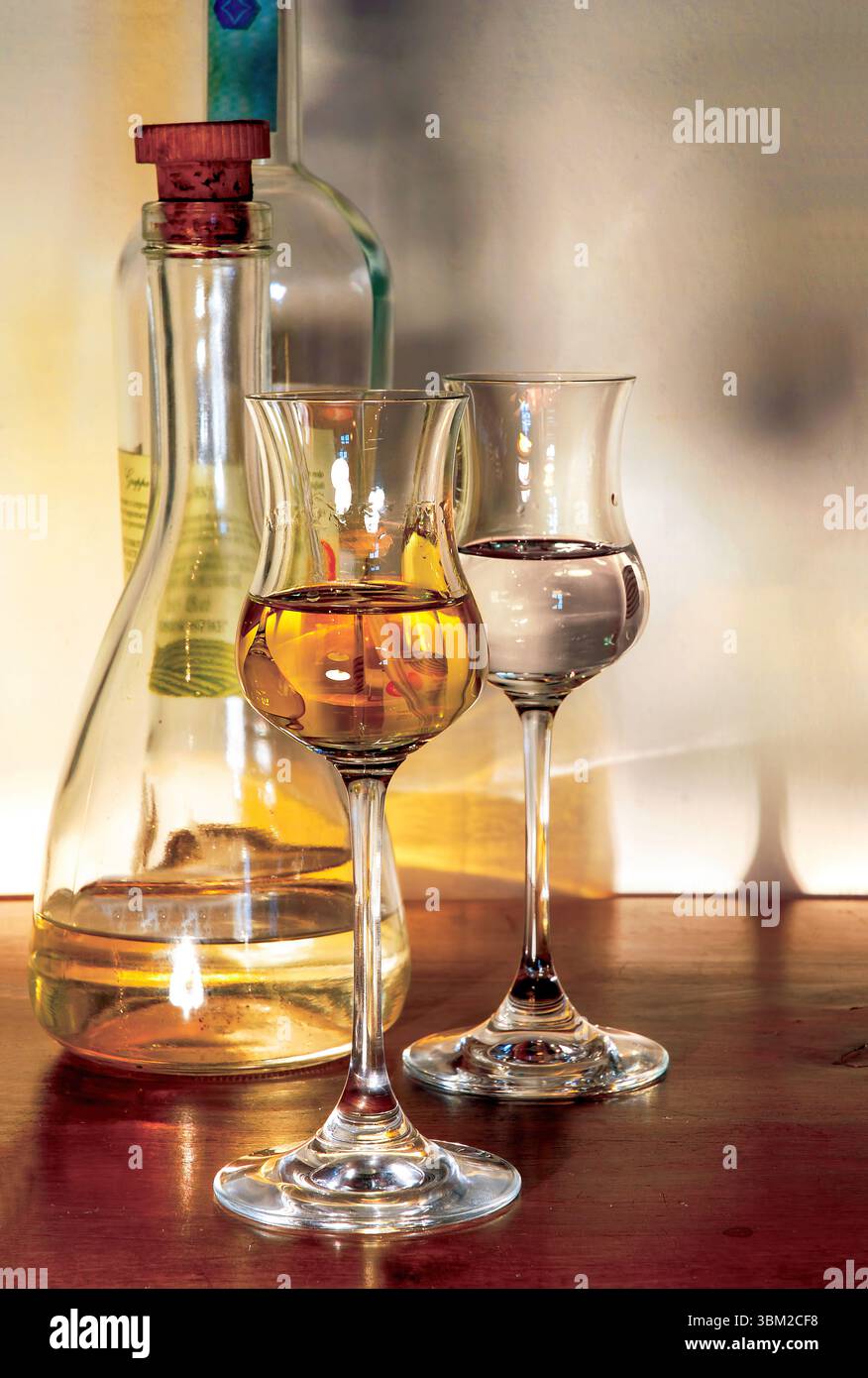 Zwei Tulpengläser mit verschiedenen Mengen goldenem Likör stehen neben einer teilweise vollen Flasche mit Korkstopfen auf einem polierten Holz Stockfoto