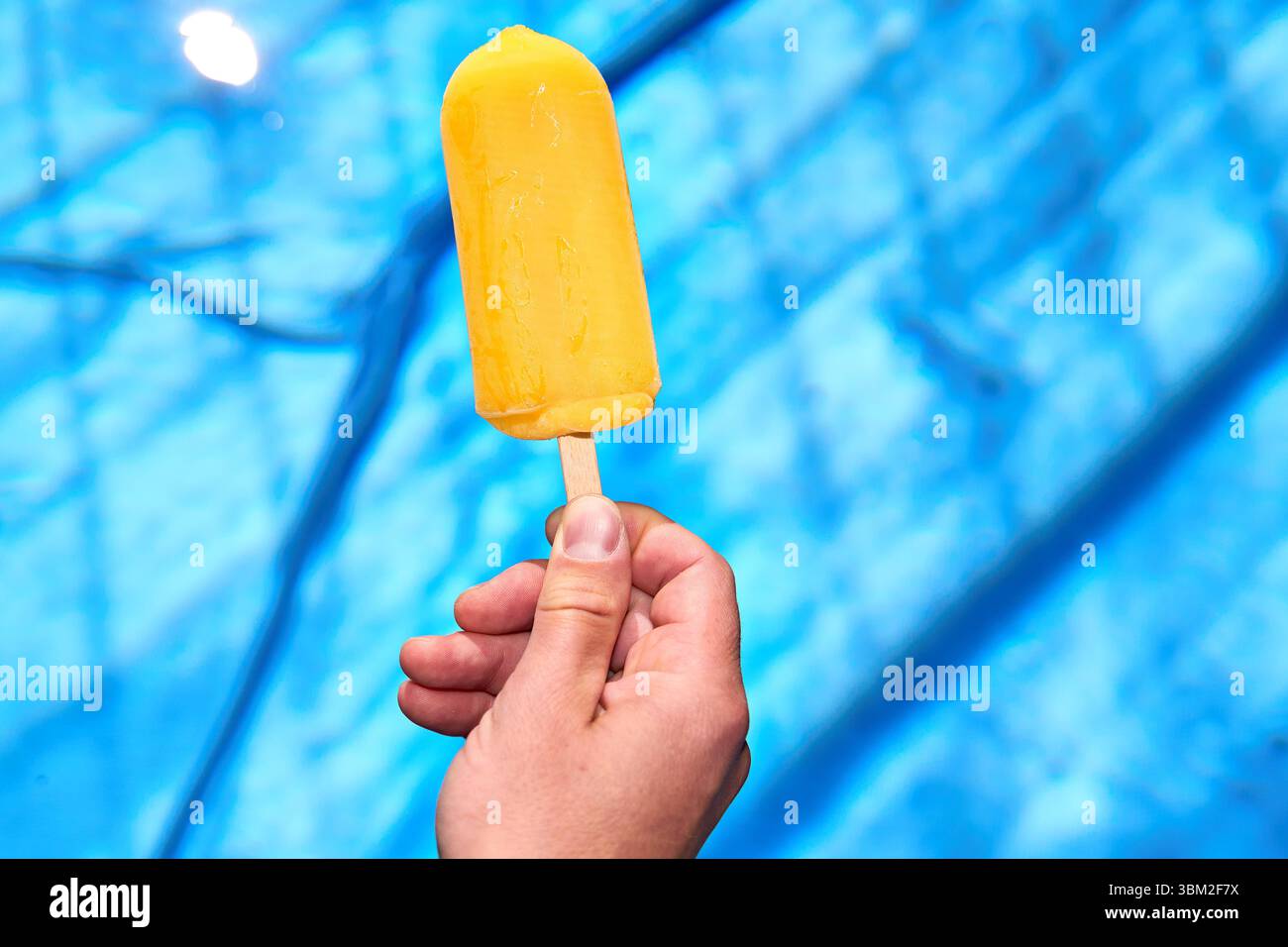 Bayern, Deutschland - 24. Juni 2025: Ein gelbes Wassereis auf einem Stock wird vor einem Schwimmbad mit ruhigem, klarem Wasser gehalten - ideal für heiße Sommertage. Das Bild symbolisiert Erfrischung, Abkühlung und den Genuss eines kühlen Imbisses im Sommer. *** Ein gelbes Wassereis am Stiel wird vor einem Swimmingpool gehalten mit ruhigem stimmel Wasser ideal für heiße Sommertage. Das Bild symbolisiert Erfrischung, Abkühlung und den Genuss eines kühlen Snacks im Sommer. Stockfoto
