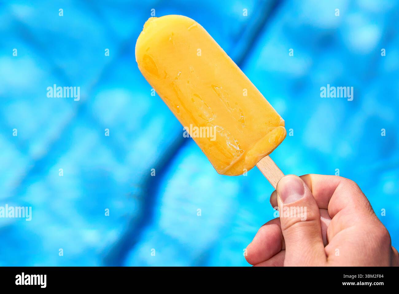 Bayern, Deutschland - 24. Juni 2025: Ein gelbes Wassereis auf einem Stock wird vor einem Schwimmbad mit ruhigem, klarem Wasser gehalten - ideal für heiße Sommertage. Das Bild symbolisiert Erfrischung, Abkühlung und den Genuss eines kühlen Imbisses im Sommer. *** Ein gelbes Wassereis am Stiel wird vor einem Swimmingpool gehalten mit ruhigem stimmel Wasser ideal für heiße Sommertage. Das Bild symbolisiert Erfrischung, Abkühlung und den Genuss eines kühlen Snacks im Sommer. Stockfoto