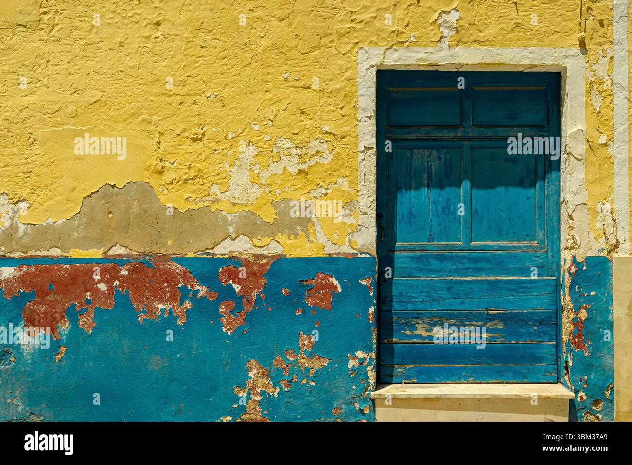 Eine alte, verwitterte Holztür in einem leuchtenden Türkisblau ist in einer Wand mit abblätternder gelber und blauer Farbe in Ciutadella, Spanien, angebracht. Stockfoto