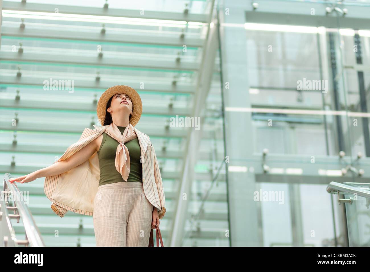 Mitten im Schuss einer weißen hübschen, eleganten Frau mit Strohhut, die nach oben blickt. Im Hintergrund befindet sich ein modernes Geschäftsgebäude. Stockfoto