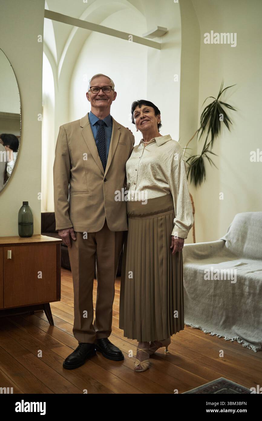 Porträt eines älteren Paares, das zusammen in einem gemütlichen Wohnzimmer mit warmer Beleuchtung steht, in formeller Kleidung für eine Party oder ein Konzert gekleidet ist, lächelt und sich liebevoll umschließt Stockfoto