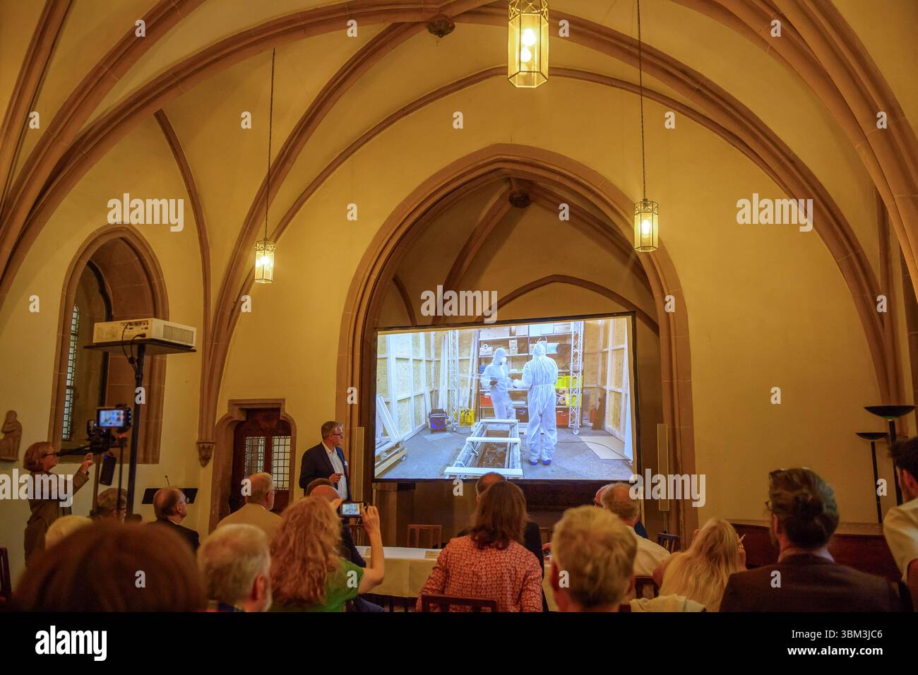 24. Juni 2025, Sachsen-Anhalt, Magdeburg: Donat Wehner (l) vom Landesamt für Denkmalpflege und Archäologie Sachsen-Anhalt kommentiert in der Sakristei des Magdeburger Doms Medienvertreter in einer Live-Videoübertragung aus dem Inneren des Gehäuses um das Grab Ottos des Großen, in dem Archäologen in Schutzanzügen am Grab Ottos des Großen zu sehen sind. Seit Januar führen das Landesamt für Denkmalpflege und Archäologie und die Kulturstiftung Sachsen-Anhalt Untersuchungen zur Erhaltung des Denkmals durch. Die Ergebnisse der Untersuchung Stockfoto