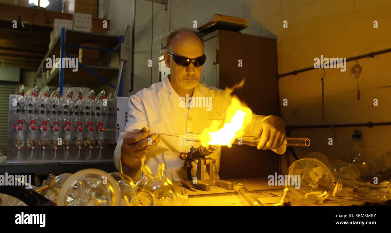 Formender männlicher Glasbläser, der die Flamme im Studio auf eine Glasröhre leitet, mit Taschenlampe und Labormantel Stockfoto