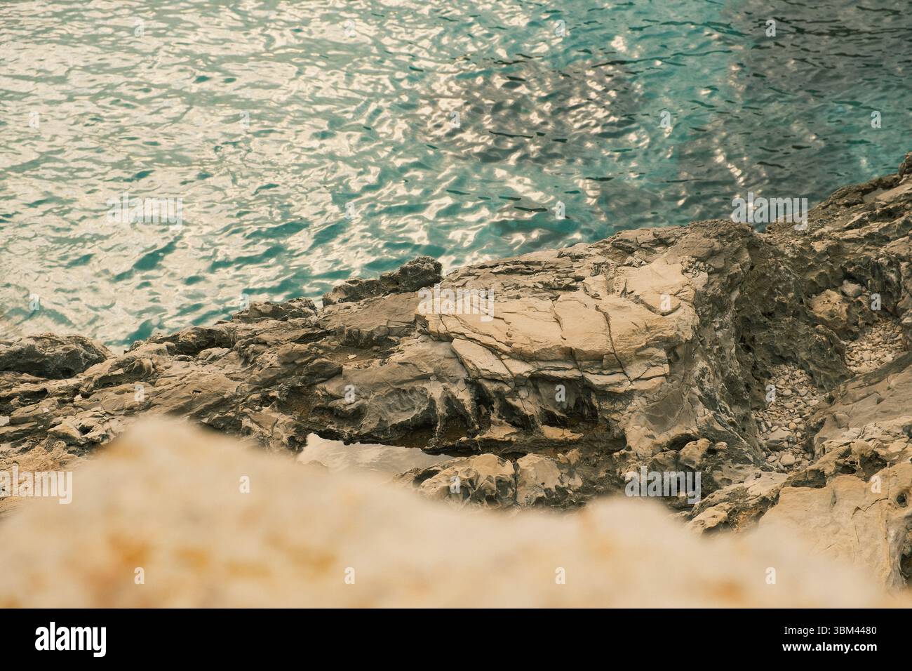 Scharfe Kalksteinklippe über türkisfarbenem Wasser der Adria. Natürliche Küstenstruktur mit zerklüfteten Felsformationen und Sonnenreflexionen auf dem Wasser. Stockfoto