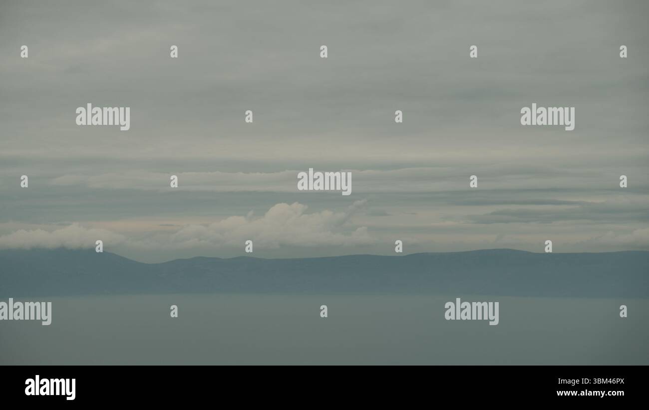 Das ruhige Wasser der Adria verschmilzt nahtlos mit den nebeligen Silhouetten der fernen Berge, die unter schweren, mehrschichtigen Wolken verborgen sind. Stockfoto