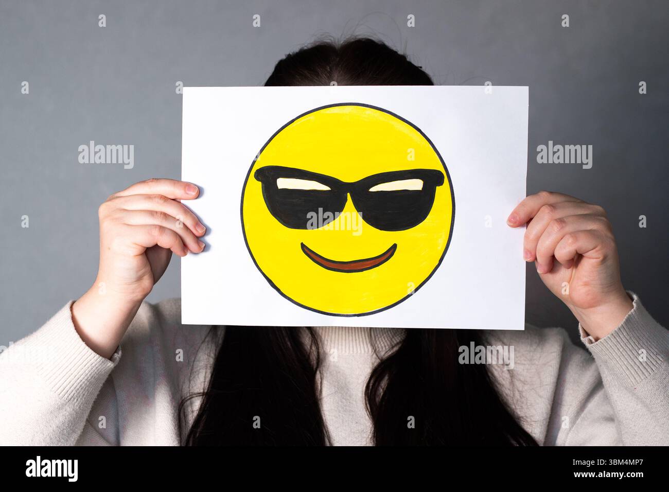 Frau, die ein glückliches Emoji mit Sonnenbrillen-Maske vor dem Gesicht hält. Social Media Trends, Gen Z Marketing, World Emoji Day 17. Juli. Stockfoto