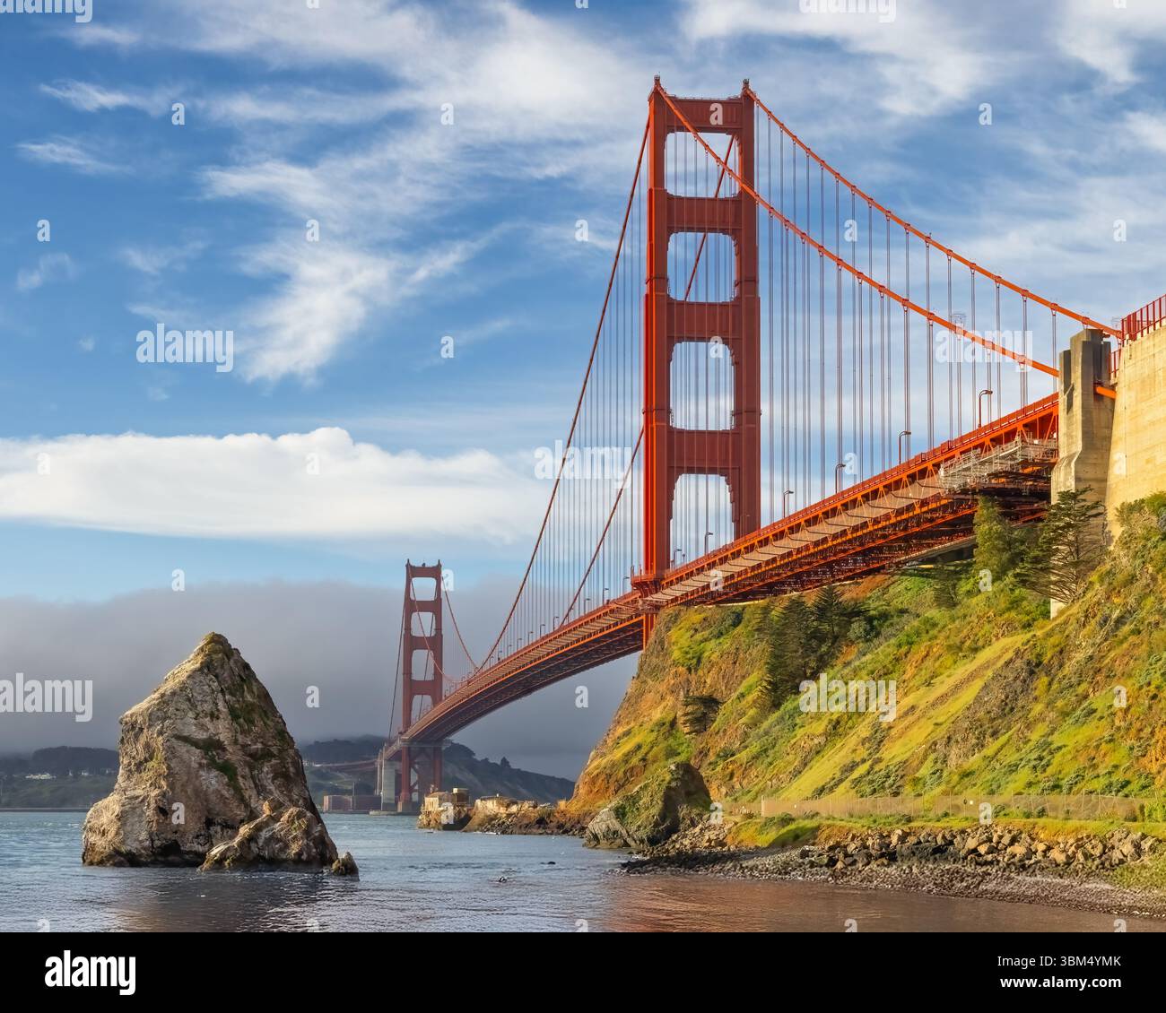 Die majestätische Golden Gate Bridge erstreckt sich über eine riesige Wasserfläche, die von einem leuchtend blauen Himmel und weichen Wolken eingerahmt wird. Der Vordergrund weist eine große Gesteinsform auf Stockfoto