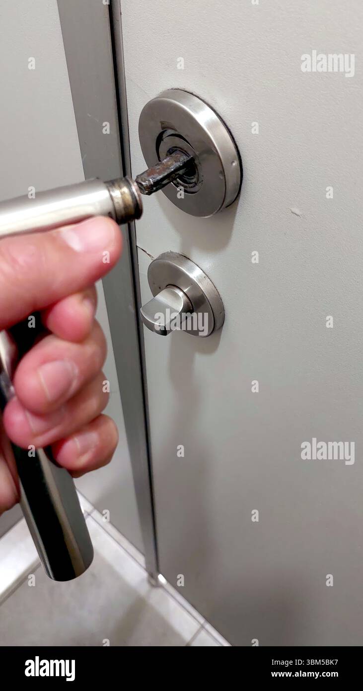 Eine Person hält einen Metallgriff, während sie an einer Tür in einem Wohnflur arbeitet. Die Tür hat ein modernes Design mit einem einfachen Schloss. Stockfoto