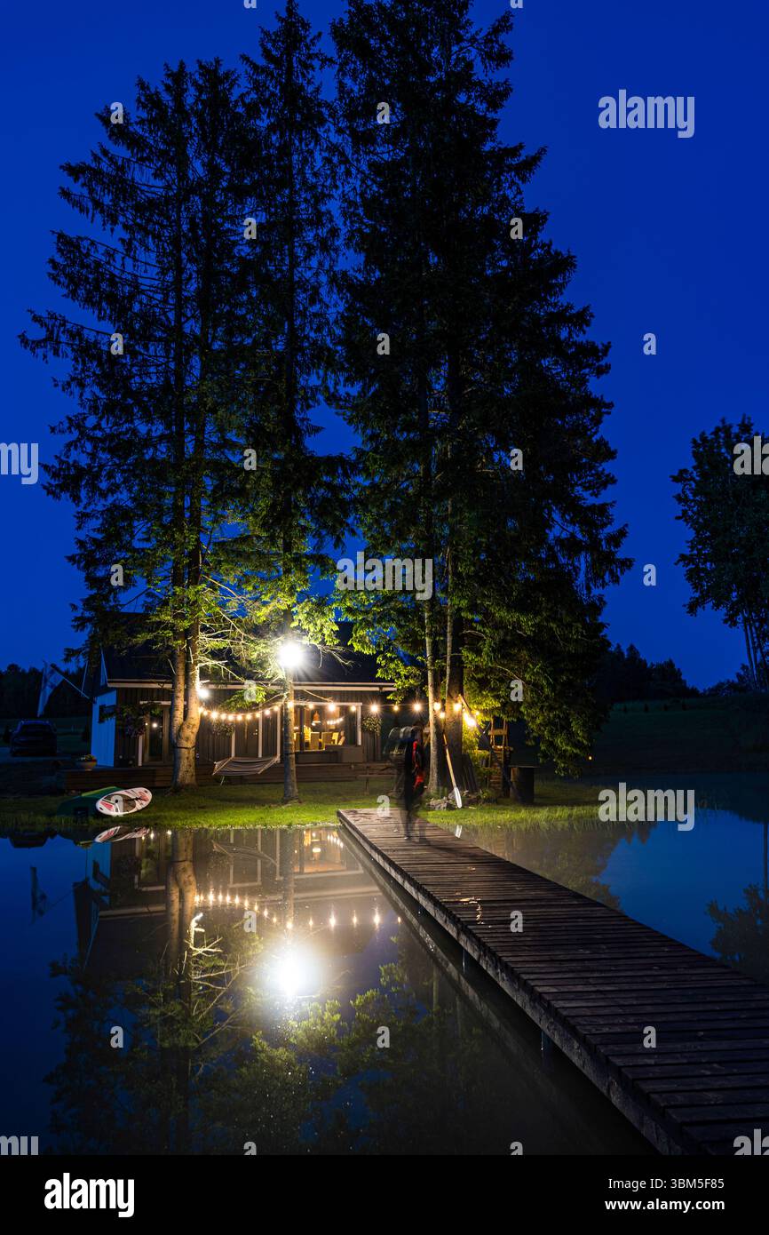 Eine gemütliche Hütte am See im ländlichen Estland, beleuchtet von warmen Außenlichtern, reflektiert in ruhigem Wasser in einer ruhigen Sommernacht. Stockfoto