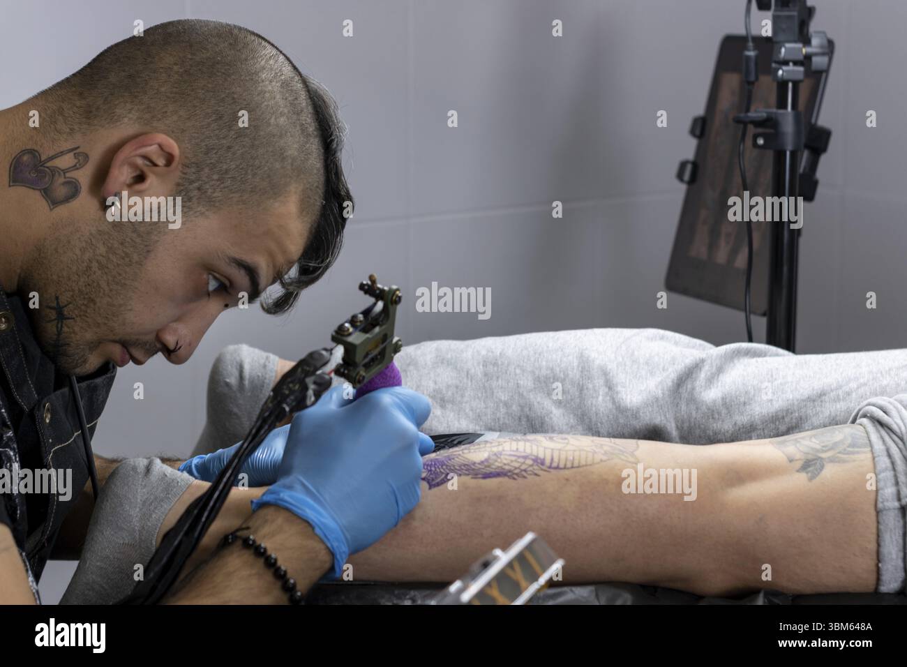 Porträt eines Tätowierers mit einem alternativen Profil bei der Arbeit, das Bein eines Kunden in seinem kleinen Atelier tätowiert. Kleine Unternehmen Stockfoto