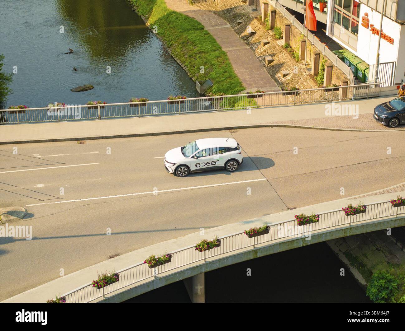 Ein weißes Auto fährt auf einer Brücke über einen Fluss, umgeben von urbaner Architektur und Natur, Deer E, Carsharing, Elektroauto, Calw, Schwarzwald Stockfoto