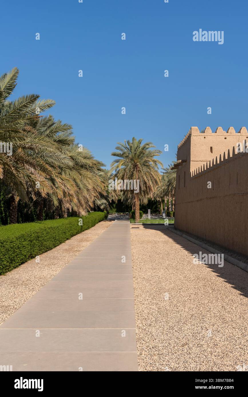 Eine lange Lehmziegelmauer mit dekorativen Zinnen und Palmen säumt einen Fußweg bei Qasr Al Muwaiji in Al Ain Vereinigte Arabische Emirate. Stockfoto