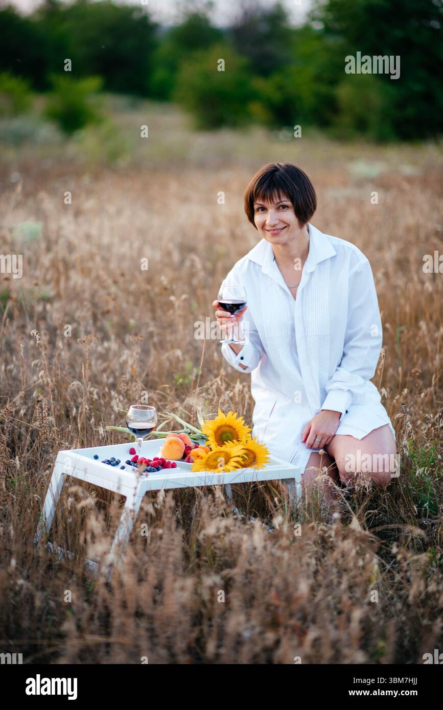 Brünette vor dem Hintergrund eines trockenen Feldes. Lächelndes Mädchen mit einem Glas Wein im Urlaub. Obst, Blumen und Wein, um Ihre Stimmung zu heben. Stockfoto