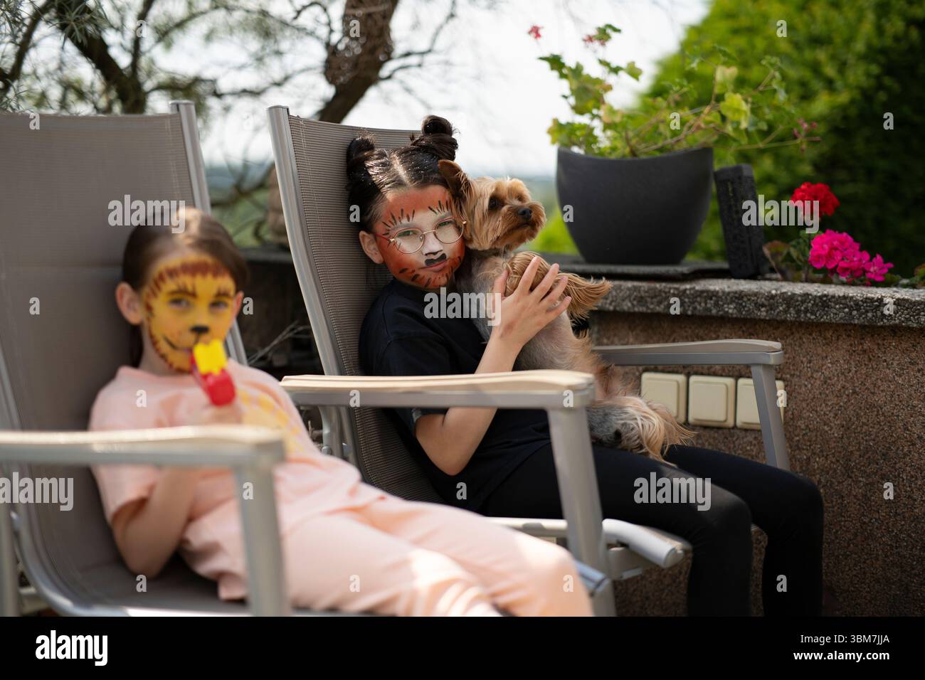 Zwei Schwestern mit tierischer Gesichtsfarbe sitzen auf Gartenstühlen in ihrem Garten. Konzept: Kinderfreude, Geschwisterbindung, Outdoor-Spaß, Fantasie, Familienmutter Stockfoto