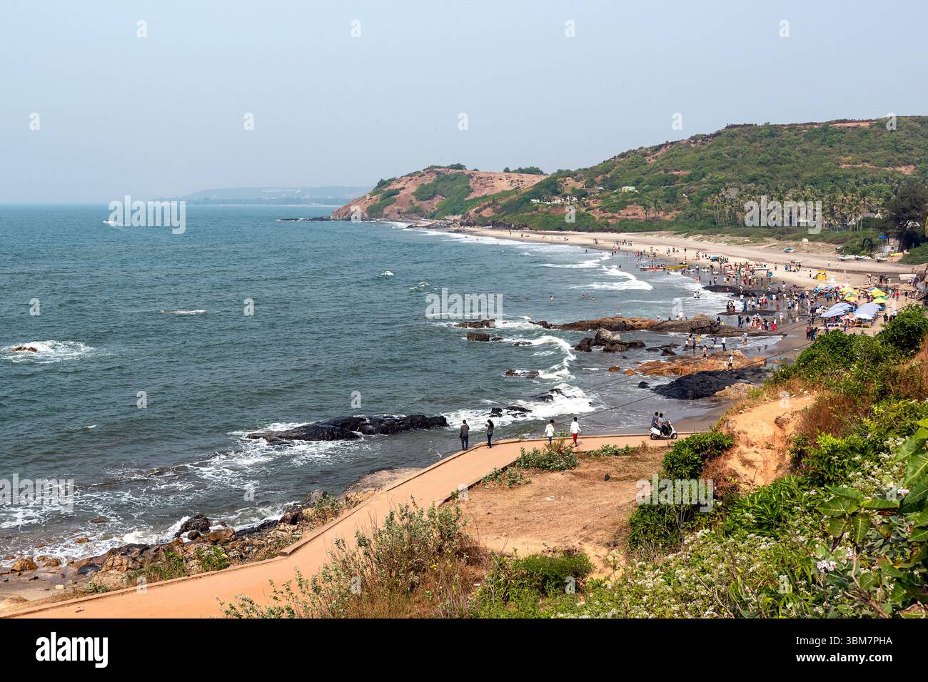 Am Anjuna Beach in Goa, Indien, treffen sanfte Wellen auf zerklüftete Felsformationen entlang der Küste, während zahlreiche Besucher an Strandaktivitäten teilnehmen Stockfoto