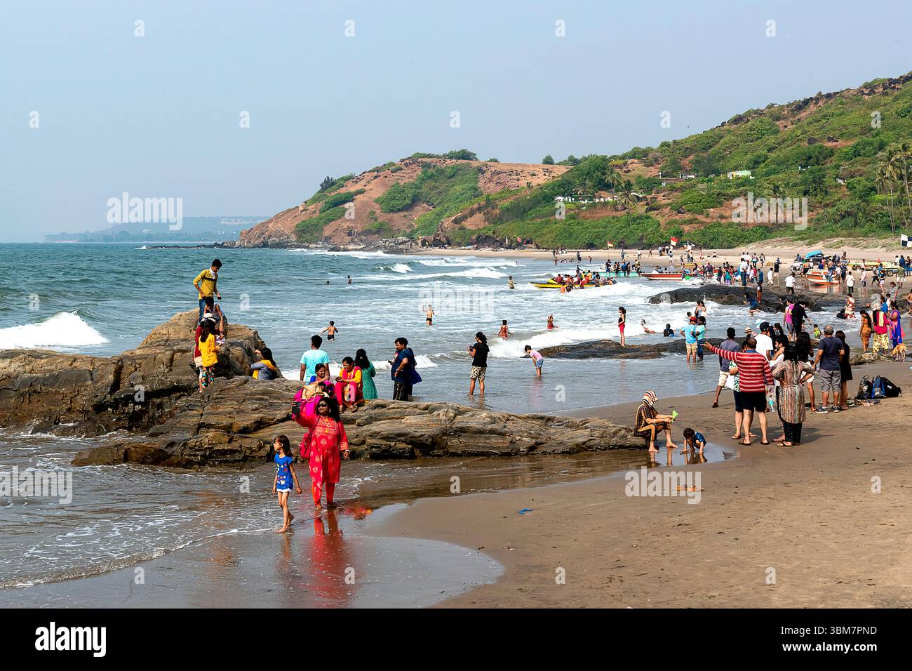 Am Anjuna Beach in Goa, Indien, treffen sanfte Wellen auf zerklüftete Felsformationen entlang der Küste, während zahlreiche Besucher an Strandaktivitäten teilnehmen Stockfoto
