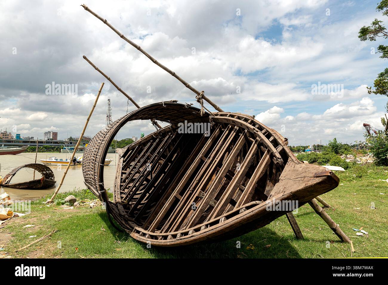 Ein verwittertes Holzboot liegt kopfüber neben einem Fluss, umgeben von Gras und Trümmern. Stockfoto