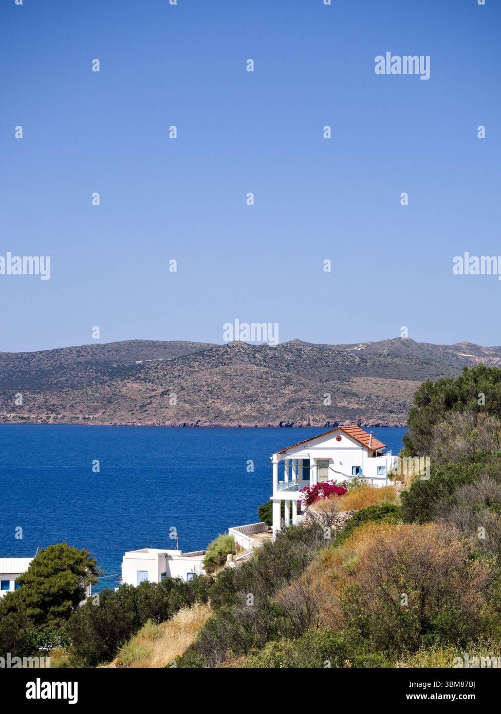 Weißes Haus auf einem Hügel an der Küste auf der Insel Milos mit Meer und Bergen hinter einem wolkenlosen blauen Himmel. Stockfoto