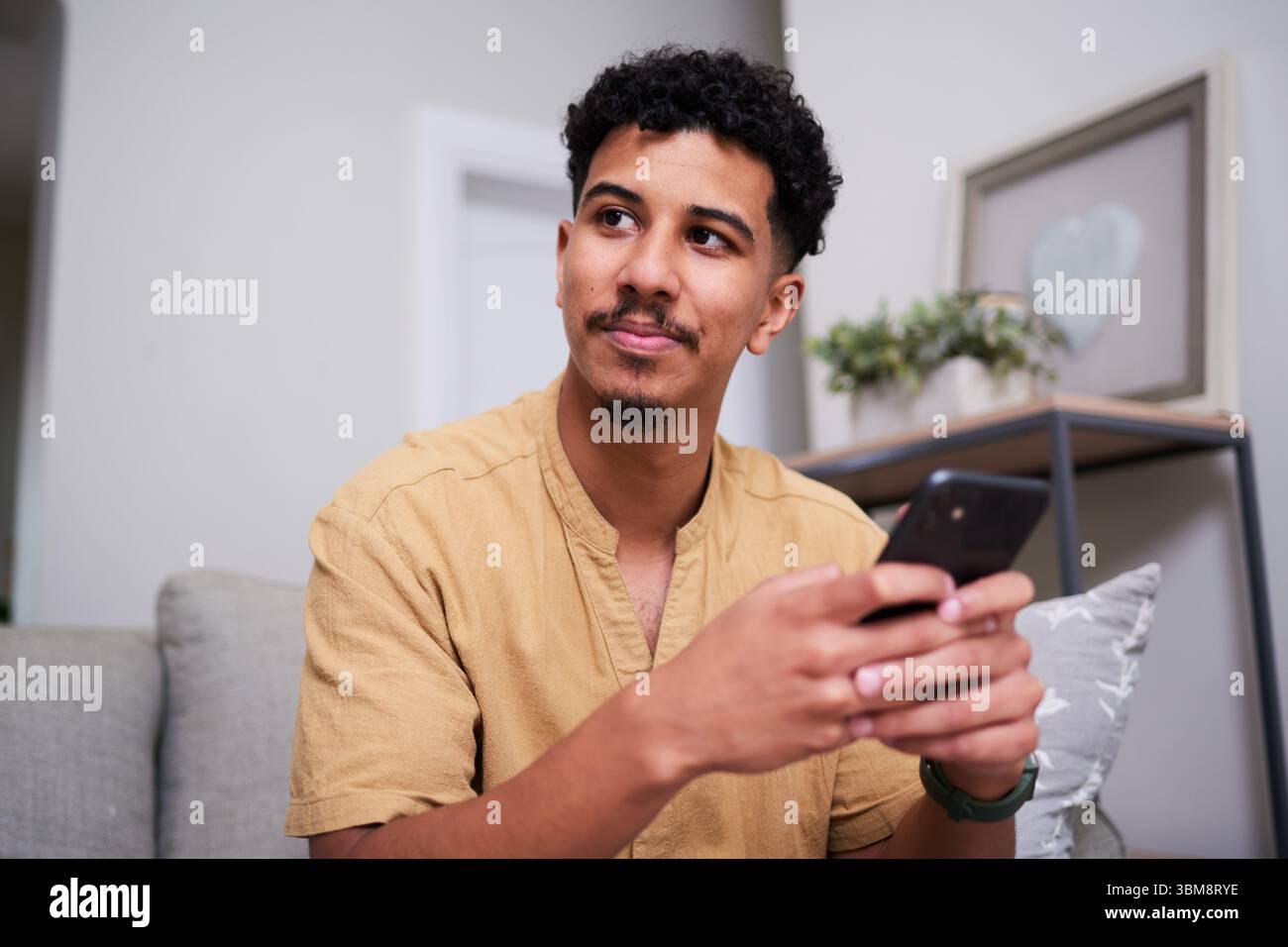 Nachdenklicher junger Erwachsener, der ein Smartphone in gemütlicher Wohnzimmeratmosphäre hält Stockfoto