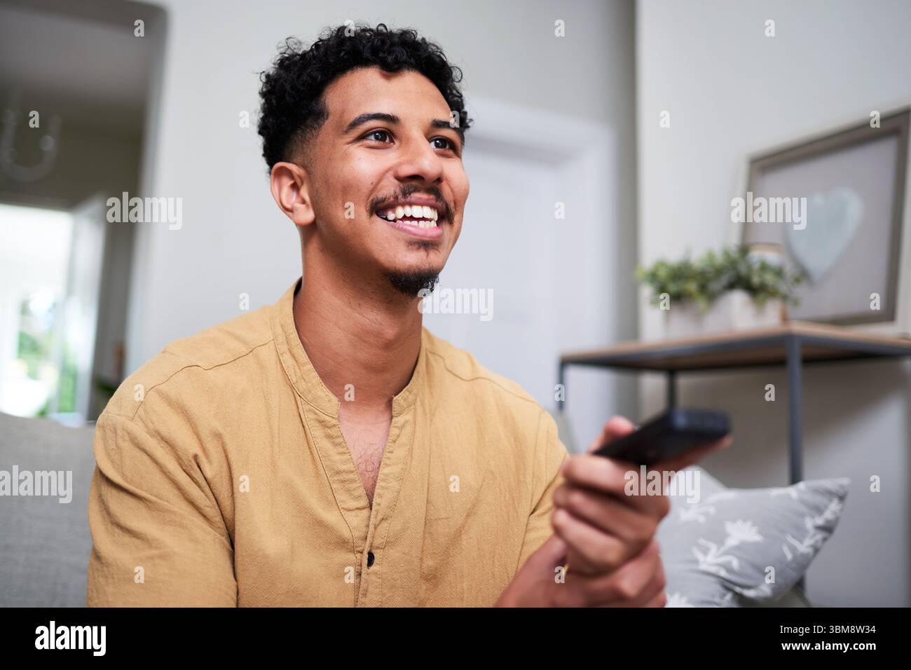 Mann lächelt beim Fernsehen in einer gemütlichen Wohnzimmerumgebung mit natürlicher Beleuchtung Stockfoto