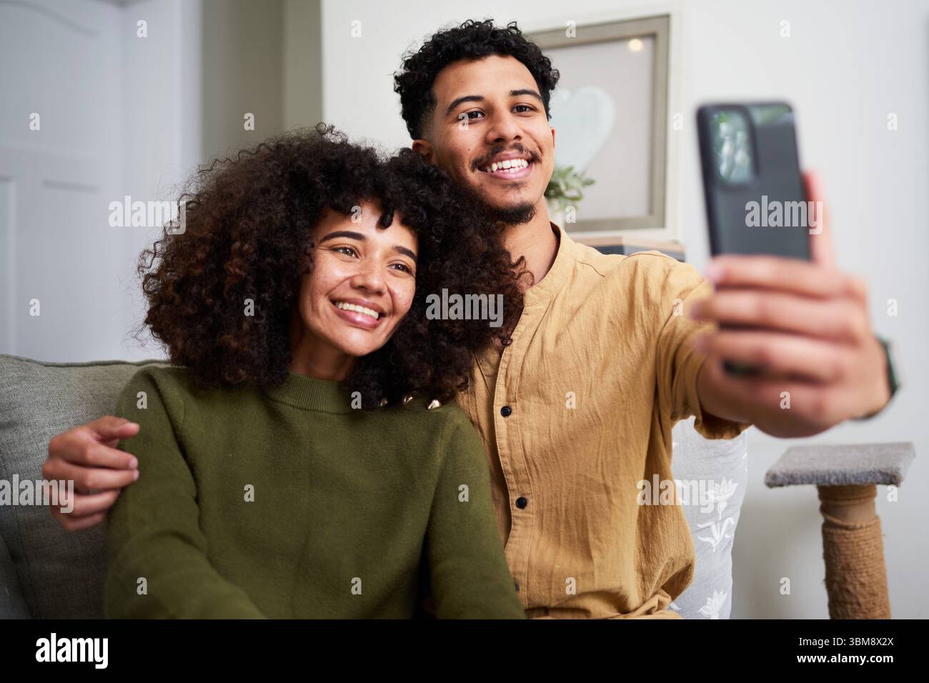 Ein lächelndes Paar sitzt zusammen und macht ein Selfie zu Hause in einem komfortablen Wohnbereich Stockfoto