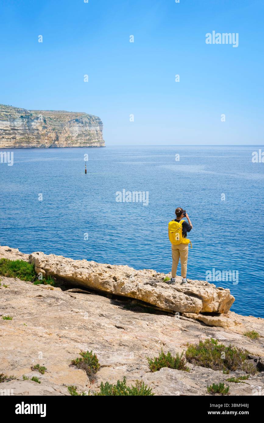 Weibliche Reisende, Rückansicht einer jungen Frau mit gelbem Rucksack, die einen Teil der Mittelmeerküste auf Malta fotografiert Stockfoto