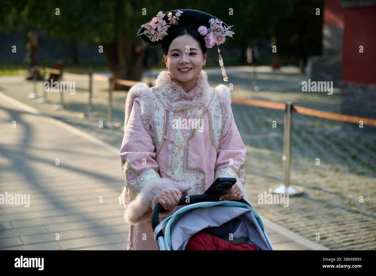 Lächelnde Frau in traditioneller chinesischer Tracht schiebt Kinderwagen im Park Stockfoto