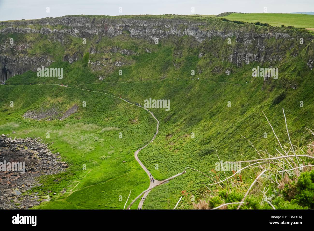 Causeway Coast Landschaft mit einem gewundenen Pfad, der einen steilen, grasbewachsenen Hügel hinunter führt, Menschen, die auf dem Weg spazieren gehen. Dramatische Naturkulisse. Stockfoto