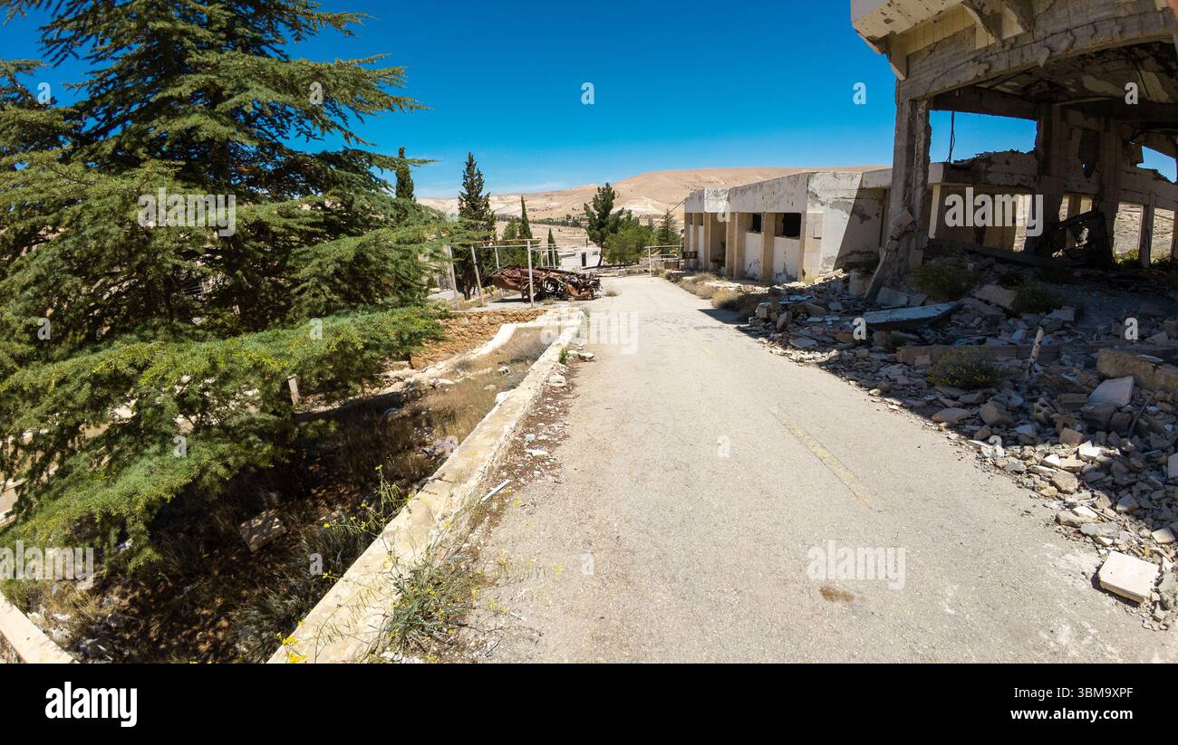 Verlassene, vom Krieg zerrissene Gebäude und Trümmer säumen eine einsame Straße unter einem klaren blauen Himmel. Stockfoto