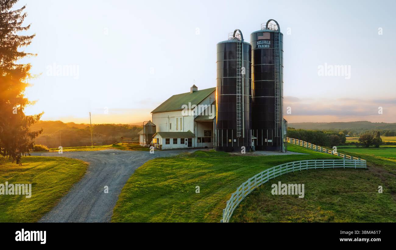 Ein ländlicher Bauernhof ist mit einer großen Scheune und zwei hoch aufragenden Silos dargestellt. Die Sonne untergeht im Hintergrund und wirft ein warmes G Stockfoto