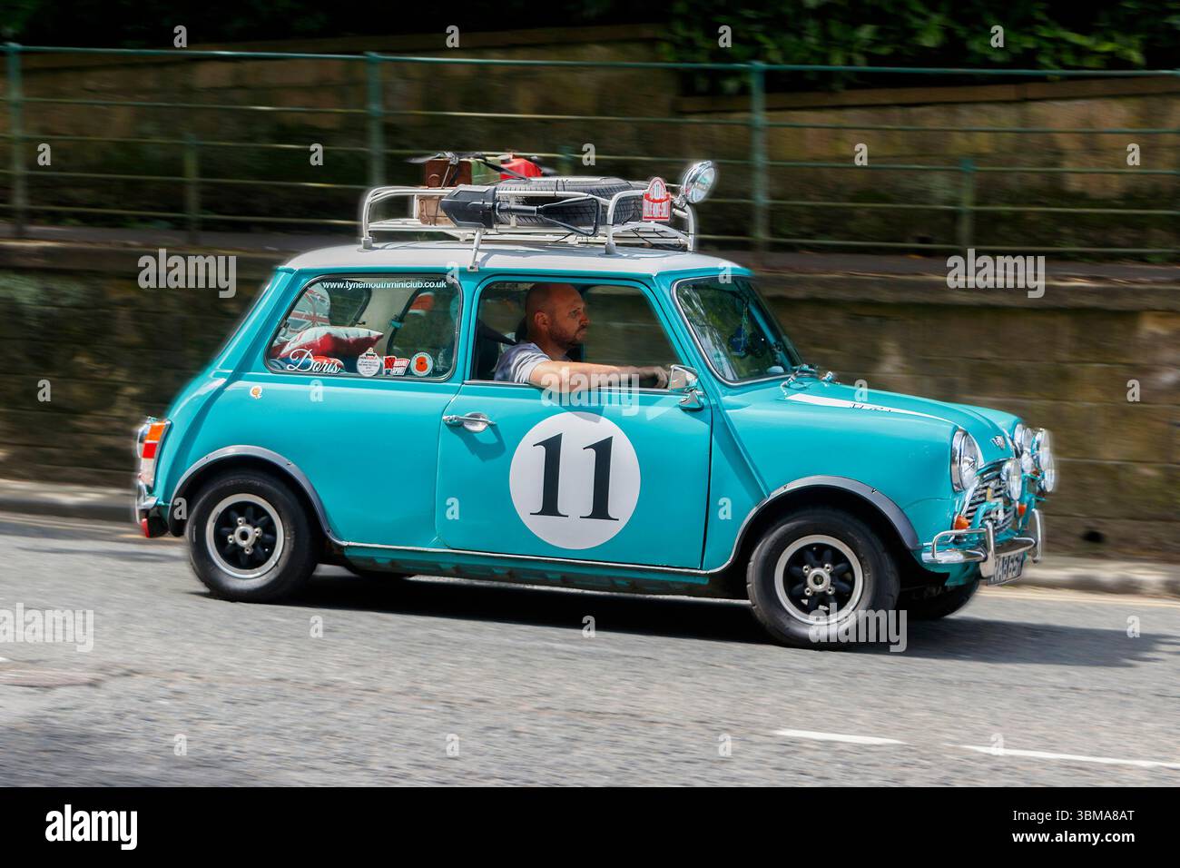 Vintage Blue Mini Cooper mit Nummer 11 Aufkleber und Dachträger, der mit einem männlichen Fahrer auf einer Stadtstraße fährt Stockfoto