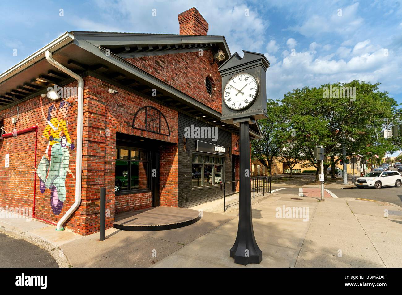 Suffern, NY - USA - 21. Juni 2025 Eine Straßenuhr steht vor der Lavagna of Suffern, einem Backsteingebäude mit einem auffälligen Wandgemälde und einer schwarzen Ladenfront an einer Ecke Stockfoto