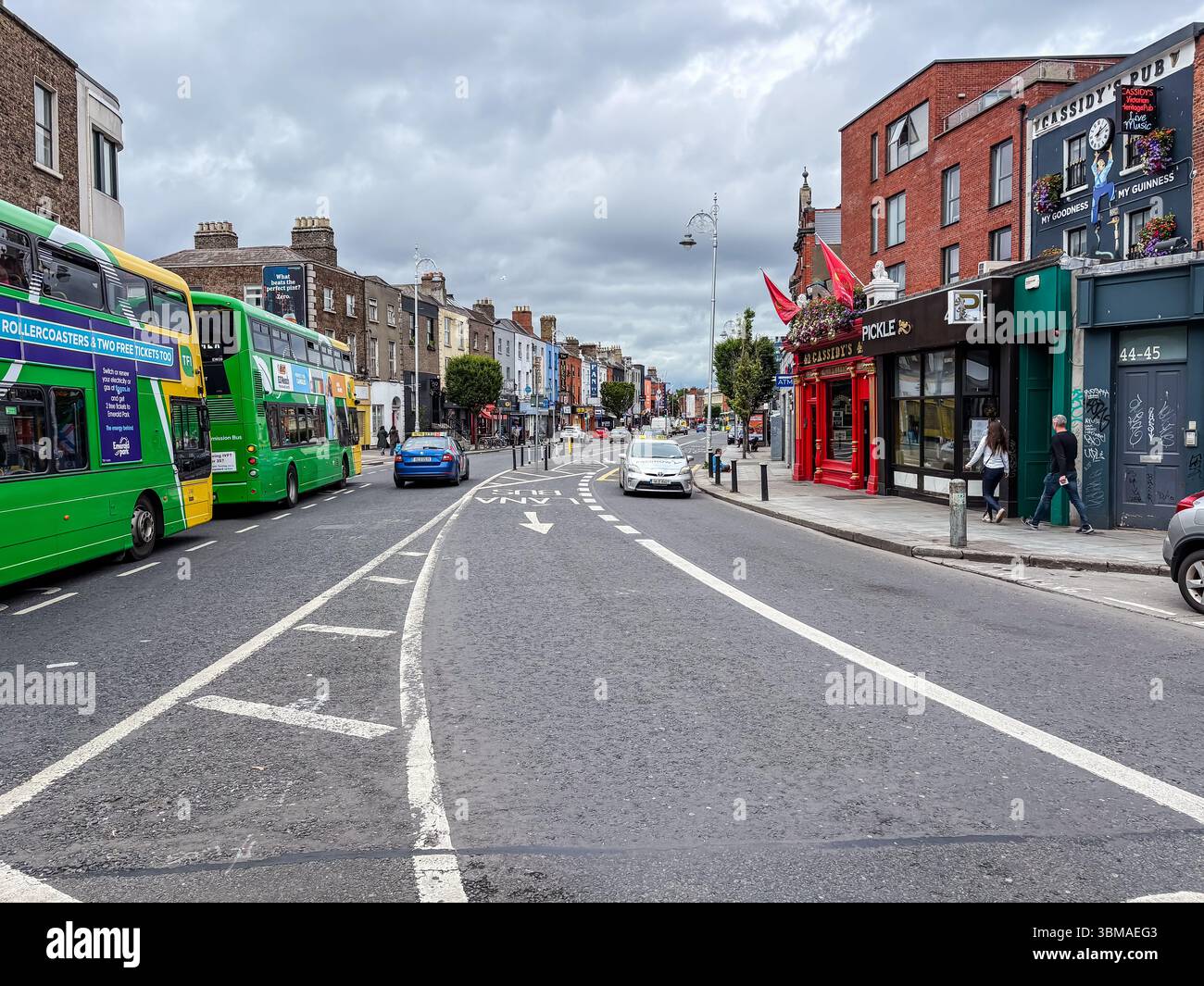 Lower Camden Street, Dublin, Irland. Belebte Straßenszene mit Geschäften, Doppeldeckerbussen, Autos und Fußgängern unter bewölktem Himmel. Stockfoto