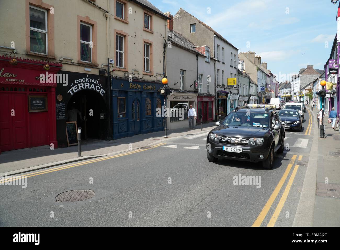 Upper Patrick St in Kilkenny, Irland mit traditionellen Ladenfronten, Pubs, Autos und Fußgängern unter blauem Himmel. Fängt den Alltag in einer historischen Geschichte ein Stockfoto