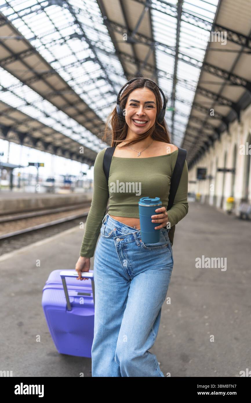 Touristen laufen im Bahnhof, ziehen Koffer und hören Musik mit Kopfhörern, halten wiederverwendbare Kaffeetasse Stockfoto