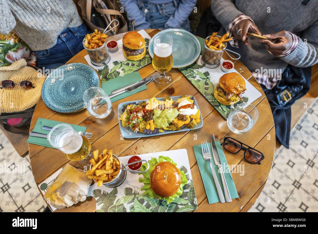 Blick von oben auf Freunde, die an einem hölzernen Restauranttisch eine Mahlzeit mit Burgern, Nachos, Pommes Frites und Getränken teilen Stockfoto