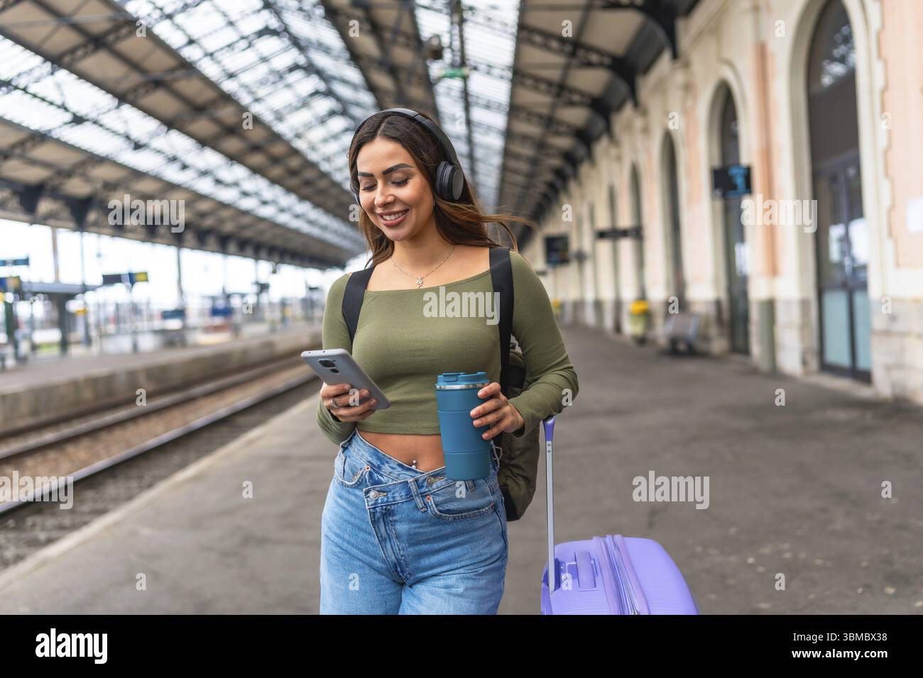 Junge Frau, die im Bahnhof mit Smartphone und Kopfhörern läuft, während sie eine wiederverwendbare Kaffeetasse und einen Rollkoffer trägt Stockfoto
