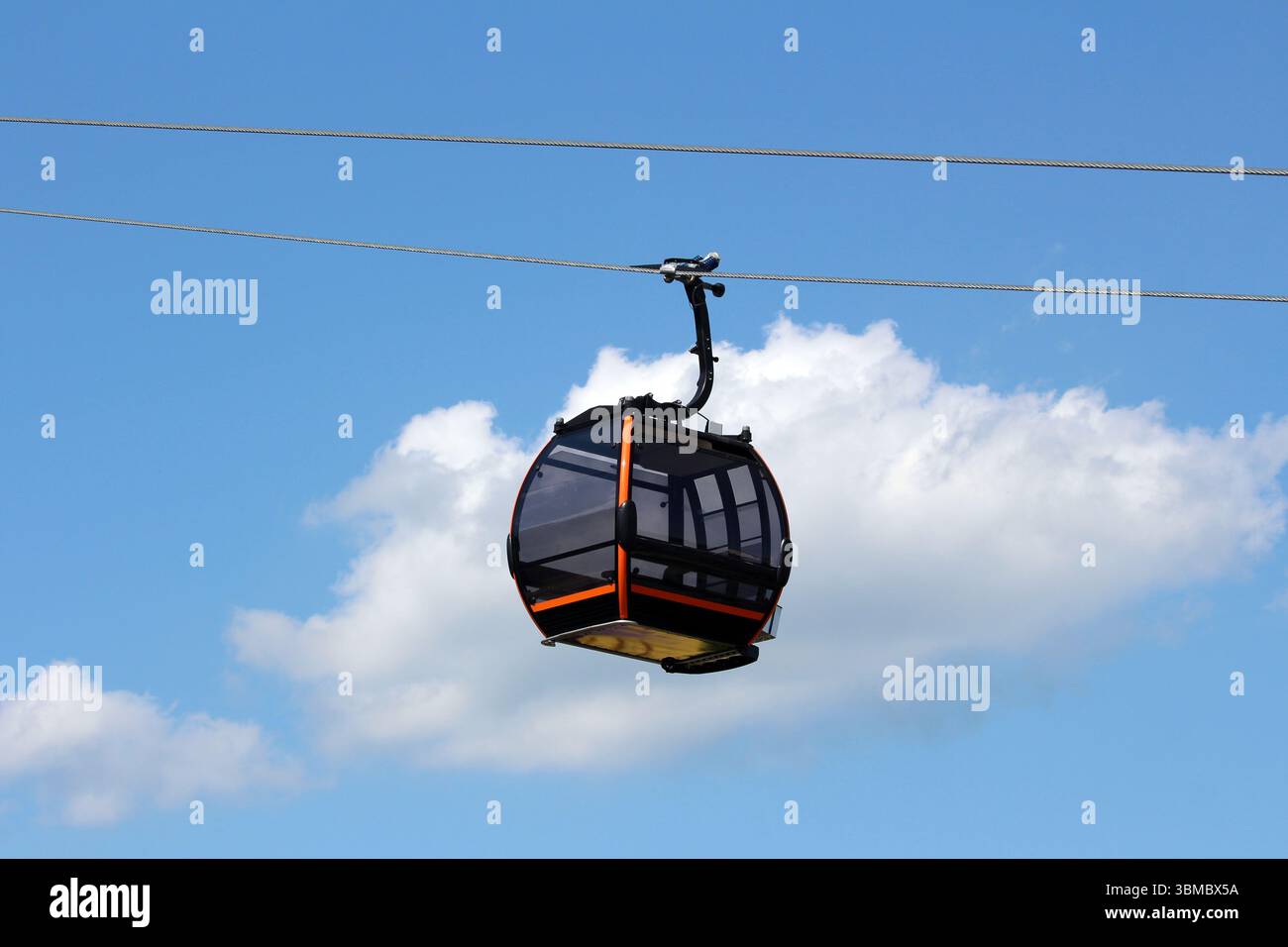 Moderne Gondelliftkabine mit getönten Fenstern und orangefarbenen Akzenten hängt an einem Stahlseil, die Silhouette vor einem hellblauen Himmel und einer weißen Wolke Stockfoto