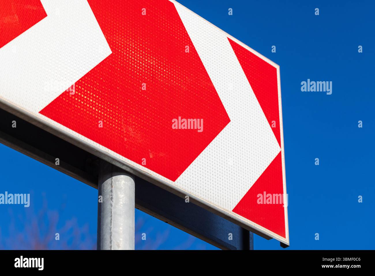 Ein rotes Richtungsschild mit nach rechts zeigenden weißen Chevrons, das vor einem leuchtend blauen Himmel steht und die Fahrtrichtung und anzeigt Stockfoto